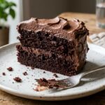 A slice of dark chocolate cake with rich chocolate buttercream frosting on a white dessert plate.